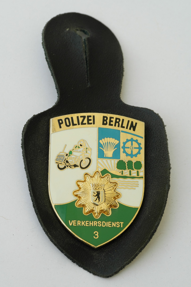 Ein schildförmiges Polizeiabzeichen mit "Polizei Berlin" in schwarzer fetter Schrift auf einem blauen Hintergrund mit einem weißen Stern und Rahmen, das auf einer Oberfläche liegt.