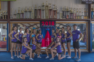 Gruppe von Cheerleadern in lila T-Shirts und schwarzen Shorts, die mit Trophäen posieren, hinter einem Banner "2018 Summit Cheerleading Champions".
