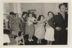 Gruppe von Menschen, die in einem Raum mit Stühlen und einem mit einer wei├čen Decke bedeckten Tisch tanzen. Im Hintergrund gibt es eine Wand mit einem Foto und Fenstern.