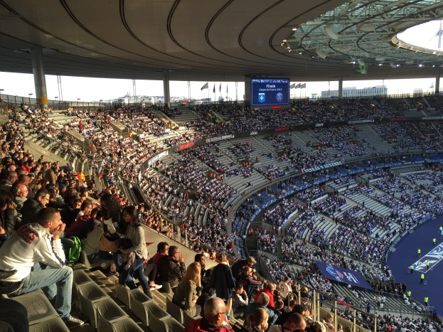 Große Menschenmenge in einem Stadion bei einem Fußballspiel, mit einer Bühne, Fahnen, Stangen, einem Bildschirm und der Allianz Arena in München, Deutschland im Hintergrund.