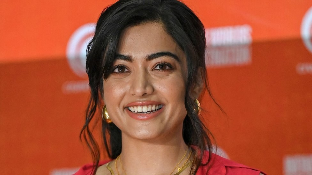 Eine Frau in einem roten Kleid mit einem eleganten Dutt, schwarzen High Heels und natürlichem Make-up lächelt in die Kamera, mit einem hellgelben Banner im Hintergrund, auf dem 'Die besten Schönheits Looks von Sonam Kapoor beim Cannes Film Festival' steht.
