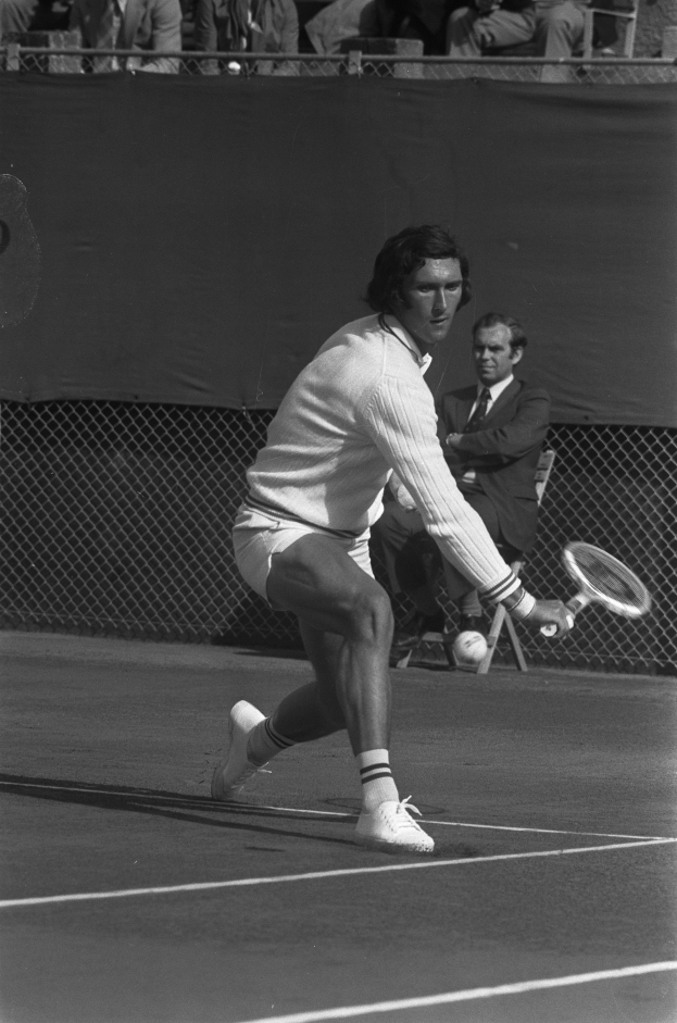 Ein Mann in Sportschuhen schlägt mit einem Tennisracket einen Ball, im Hintergrund ist ein Netz und Zuschauer auf Stühlen zu sehen.