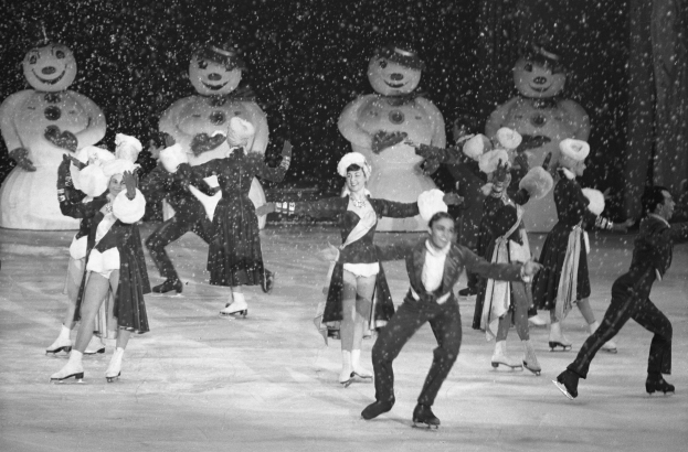Eine Gruppe von Menschen in Kostümen und Eislaufschuhen fährt auf einer Eisbahn im Schnee Schlittschuh, mit Schneemännern im Hintergrund, in einem Schwarz-Weiß-Bild.