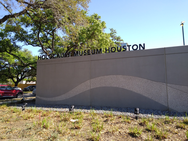 Außenansicht des Houston Holocaust Museums mit einer strukturierten Wand, verstreuten Steinen, Grünflächen, Bäumen, vorbeifahrenden Fahrzeugen, einem Straßenpfahl und einem bewölkten Himmel.