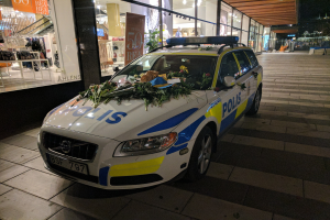 Ein mit Blumen und Blättern geschmückter Polizeiwagen steht nachts vor einem Geschäft mit Glaswänden, das Schaufensterpuppen in Kleidern und beleuchtete Schilder zeigt, mit Bäumen und Gebäuden im Hintergrund.