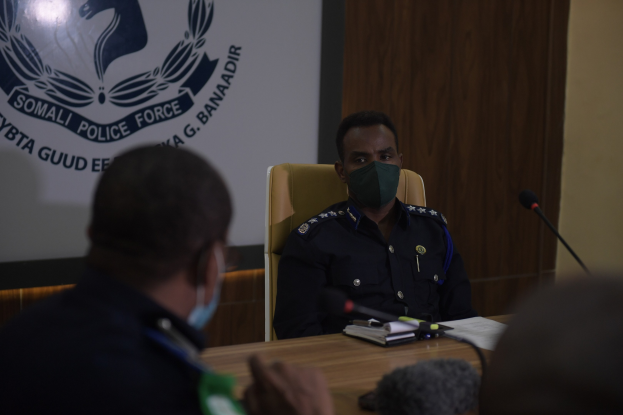 Zwei Polizeibeamte mit Masken sitzen an einem Tisch mit Mikrofonen, Büchern und anderen Gegenständen; im Hintergrund ist ein Bildschirm mit Text und einem Emblem zu sehen.