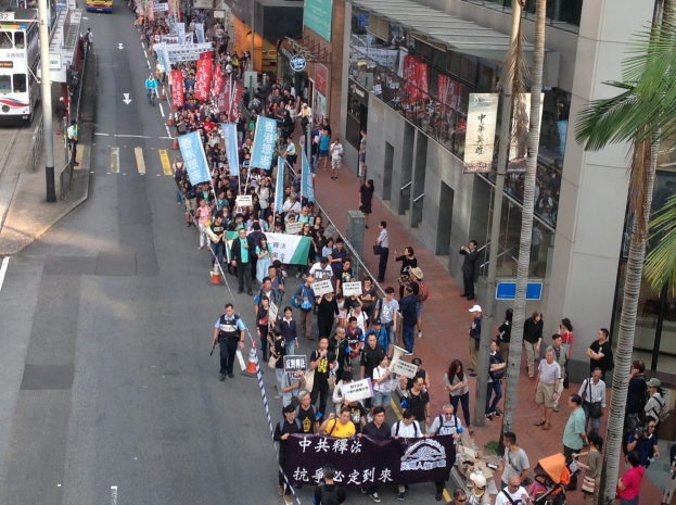 Eine große Gruppe von Menschen marschiert auf einer Straße in Hong Kong, hält Schilder und Plakate hoch, mit Bäumen, glasverkleideten Gebäuden, Fahrzeugen und Hinweistafeln im Hintergrund.