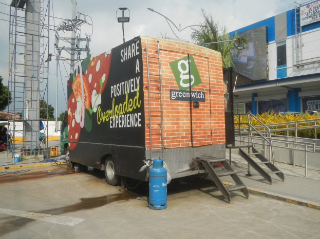 Ein Lkw, der vor einem Gebäude mit einer Tafel parkt, die "Teilen Sie ein positiv überladenes Erlebnis" lautet, Treppen, die zum Lkw führen, ein Zylinder auf dem Boden, Geländer, Strommasten mit Drähten, Laternen, Bäume und ein bewölkter Himmel im Hintergrund.