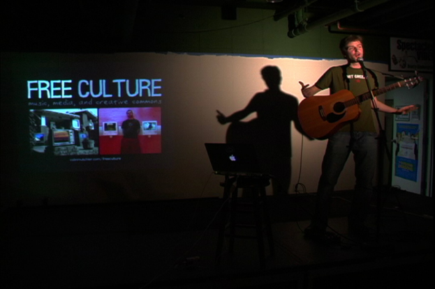 Eine Person, die eine Gitarre hält und auf einem Tisch ein Laptop und im Hintergrund ein Projektionsbildschirm mit der Aufschrift "Free Culture" zu sehen ist.