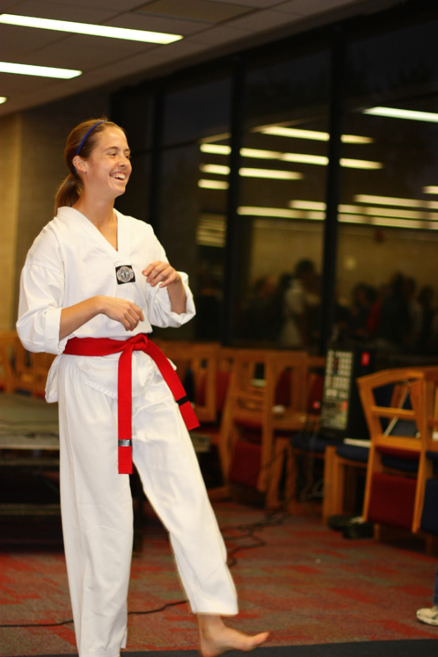 Ein Mädchen in einem Karate-Outfit mit einem roten Gürtel lacht, schaut auf etwas, mit Stühlen, einer Musikanlage und einigen Menschen dahinter und einer Wand mit Lichtern im Hintergrund.