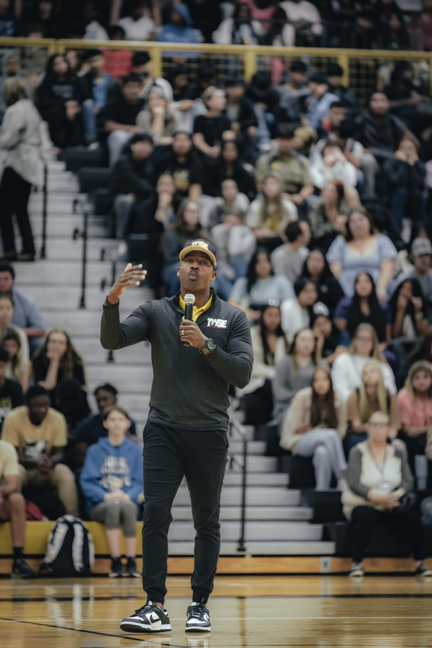 Ein Mann mit Mütze hält ein Mikrofon auf einem Basketballfeld, mit einer Gruppe von Menschen auf der Treppe und Taschen auf dem Boden dahinter.