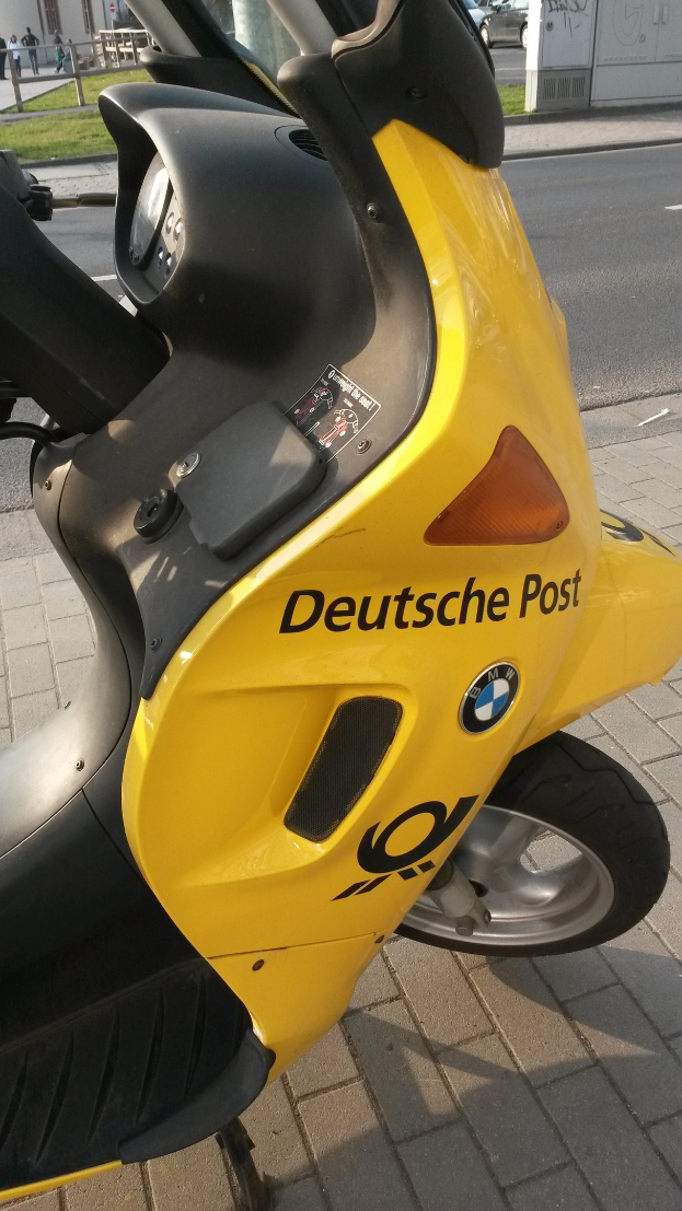 Ein gelber E-Scooter auf einer Straße mit einem sichtbaren Kennzeichen, umgeben von Gebäuden, Bäumen, Pfählen, Brettern und anderen Fahrzeugen.