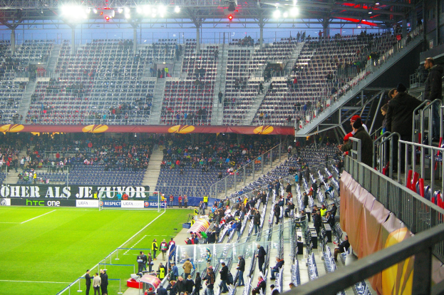 Großes Stadion voller Zuschauer bei einem Fußballspiel, mit sitzenden und stehenden Zuschauern, einer Absperrung, einem Banner auf der rechten Seite und einem leeren Abschnitt im Hintergrund mit Deckenbeleuchtung.