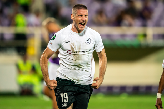 Ein Mann in einem weißen Hemd und schwarzen Shorts rennt auf einem Fussballfeld, feiert ein Tor und ist von einer Gruppe Menschen umgeben.