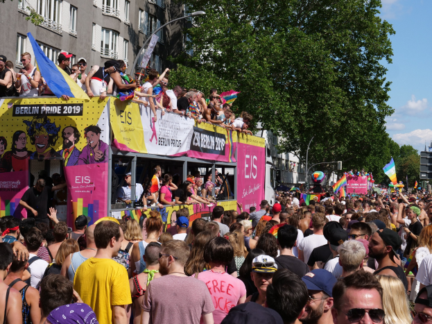 Eine große Menschenmenge, die auf der Straße in Berlin Pride 2019 marschiert, viele mit Mützen und Schutzbrillen, Fahnen haltend, mit Bannern und Gebäuden im Hintergrund bei bewölktem Himmel.