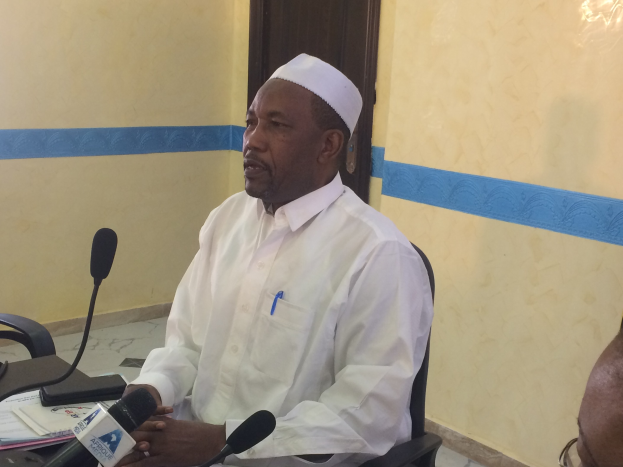 Ein Mann in einem weißen Kleid und einer Mütze sitzt an einem Tisch mit Mikrofonen, während er spricht und Papiere und Gegenstände auf dem Tisch sowie eine Person mit Brille rechts von ihm in einem Raum mit einer Tür im Hintergrund zu sehen sind.