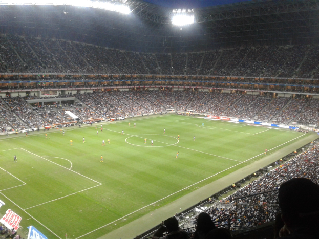 Ein großes, hell erleuchtetes Stadion voller Zuschauer, die während eines Fußballspiels jubeln und von Werbetafeln umgeben sind.