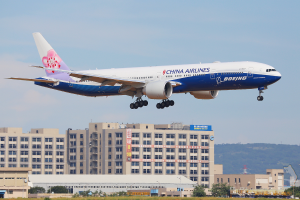 Ein China-Airlines-Boeing-777-300ER-Flugzeug startet vom Beijing-Capital-International-Airport, umgeben von grünem Gras, Bäumen, Gebäuden und einem fernen Hügel unter einem bewölkten Himmel.