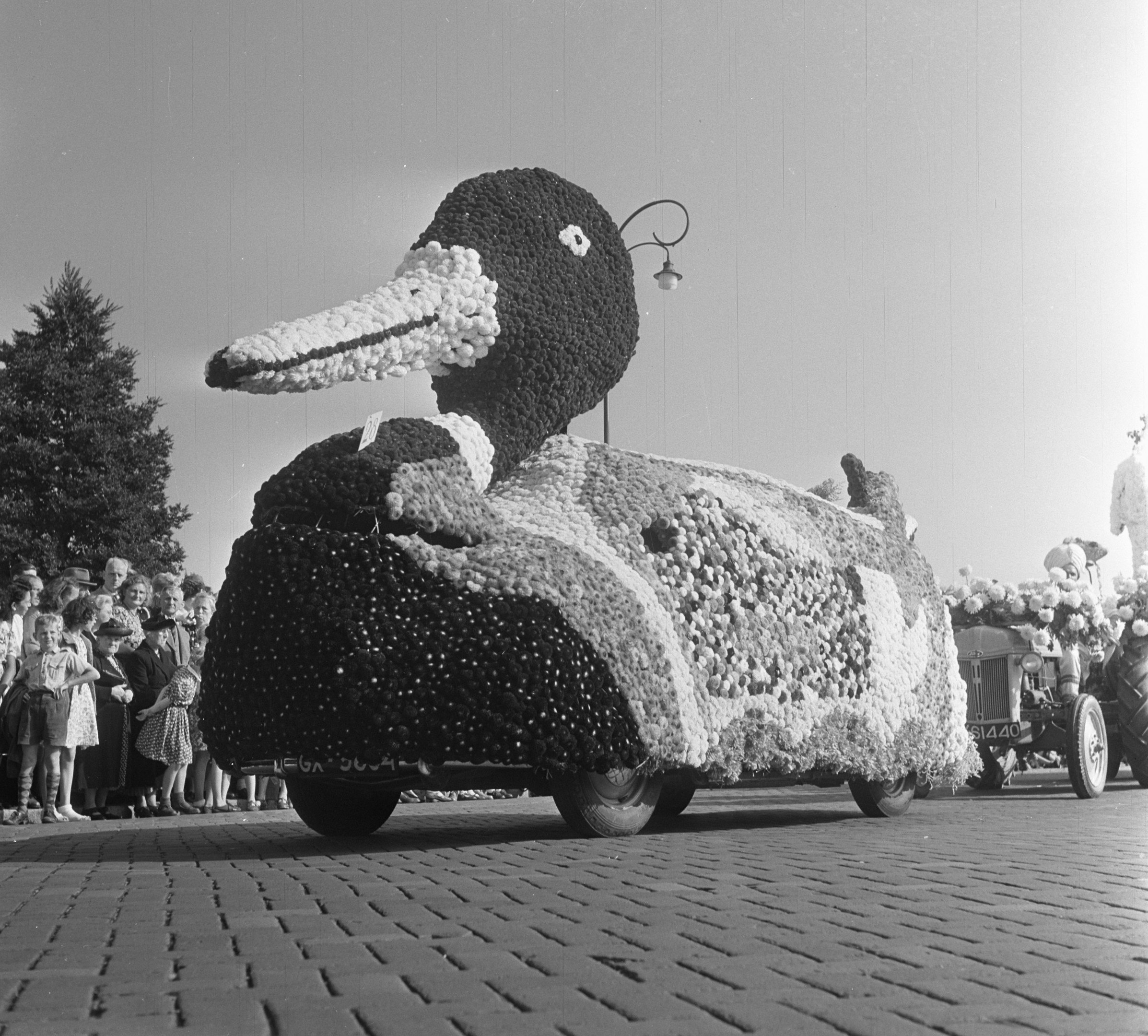 Schwarzes und weißes Foto eines Paradewagens in Form einer Ente, dekoriert mit Blumen, mit Menschen zu seiner Linken und Bäumen im Hintergrund bei klarem Himmel.