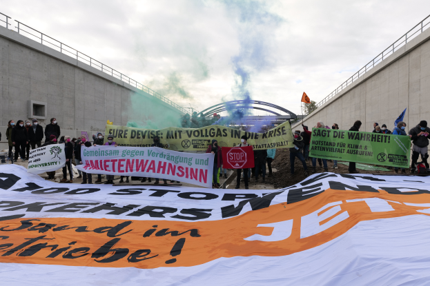 Eine Gruppe von Menschen steht vor einer großen Fahne mit der Aufschrift "Klima-Protest in Berlin", haltend Schilder und Fahnen, mit Rauch im Hintergrund und einem bewölkten Himmel darüber.