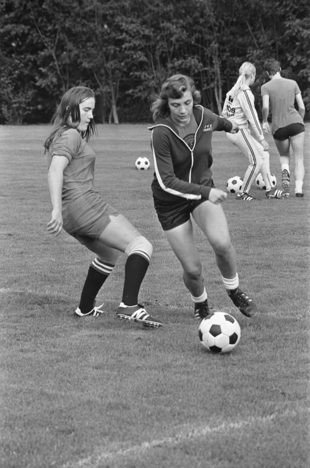 Eine Gruppe junger Frauen, die auf einem von Bäumen umgebenen Feld Fußball spielen, tragen Fußballtrikots und Turnschuhe, in Schwarz-Weiß.