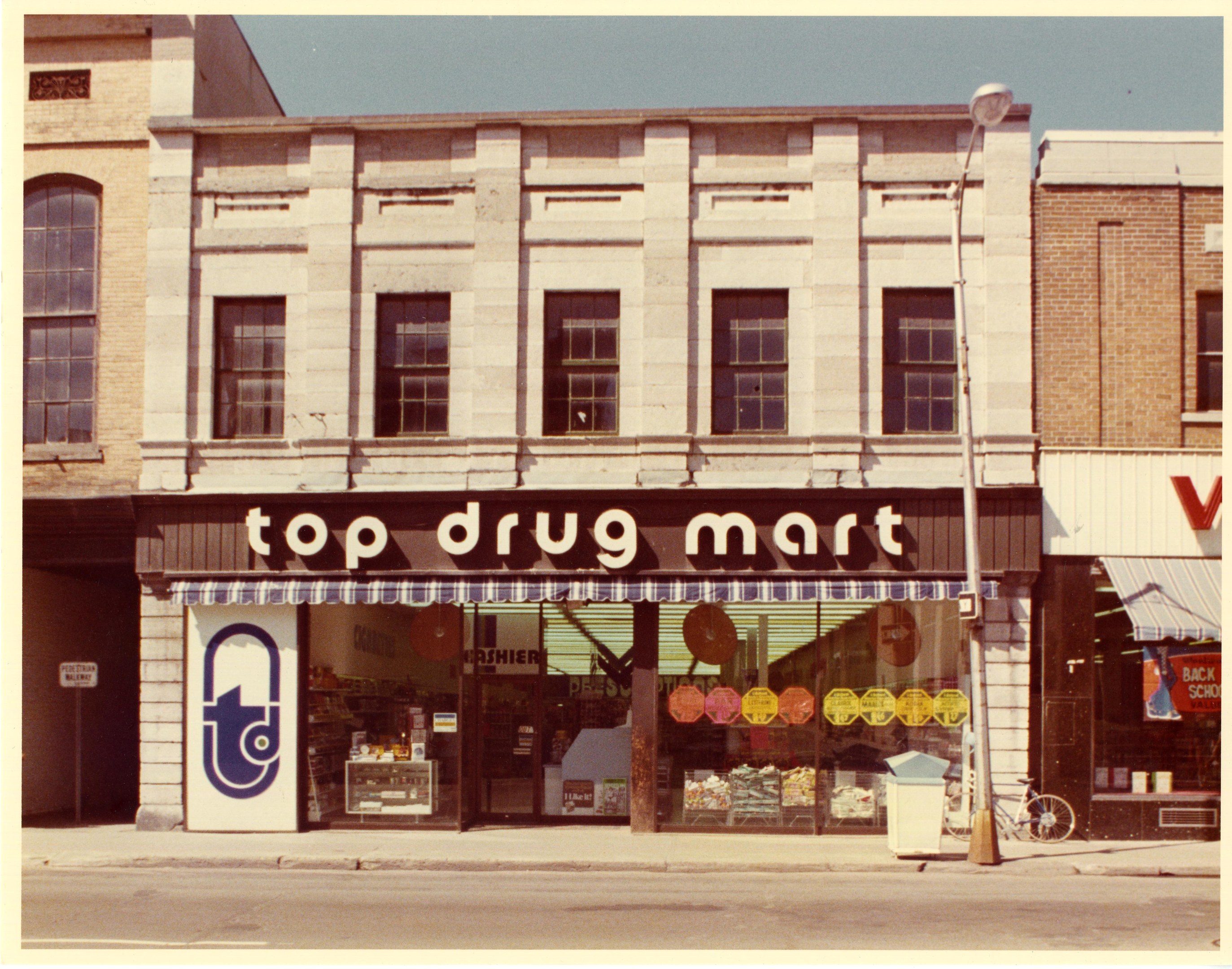 Ein Drogengeschäft an einer Straßenecke mit einem Gebäude, einem Schild, einem Straßenpfosten, einem geparkten Fahrrad, einem Müllcontainer und einem bewölkten Himmel.