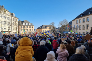 Große Menschenmenge vor einem Gebäude protestierend, mit Schildern und Fahnen, Bäumen und mehrfenstrigen Gebäuden im Hintergrund bei klarem Himmel.