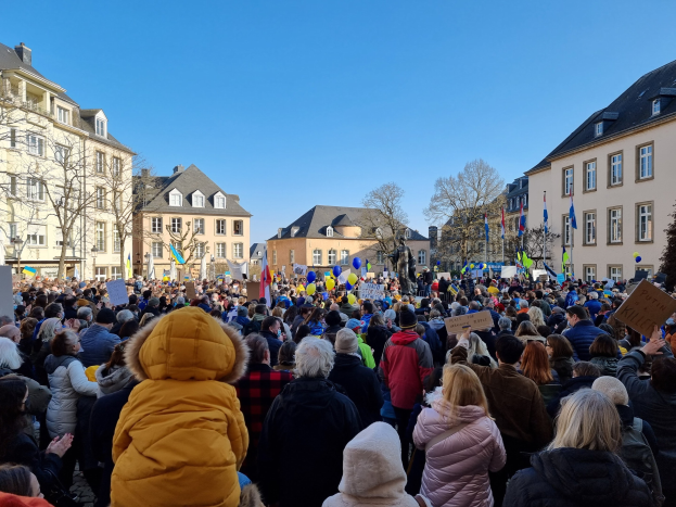 Große Menschenmenge vor einem Gebäude protestierend, mit Schildern und Fahnen, Bäumen und mehrfenstrigen Gebäuden im Hintergrund bei klarem Himmel.
