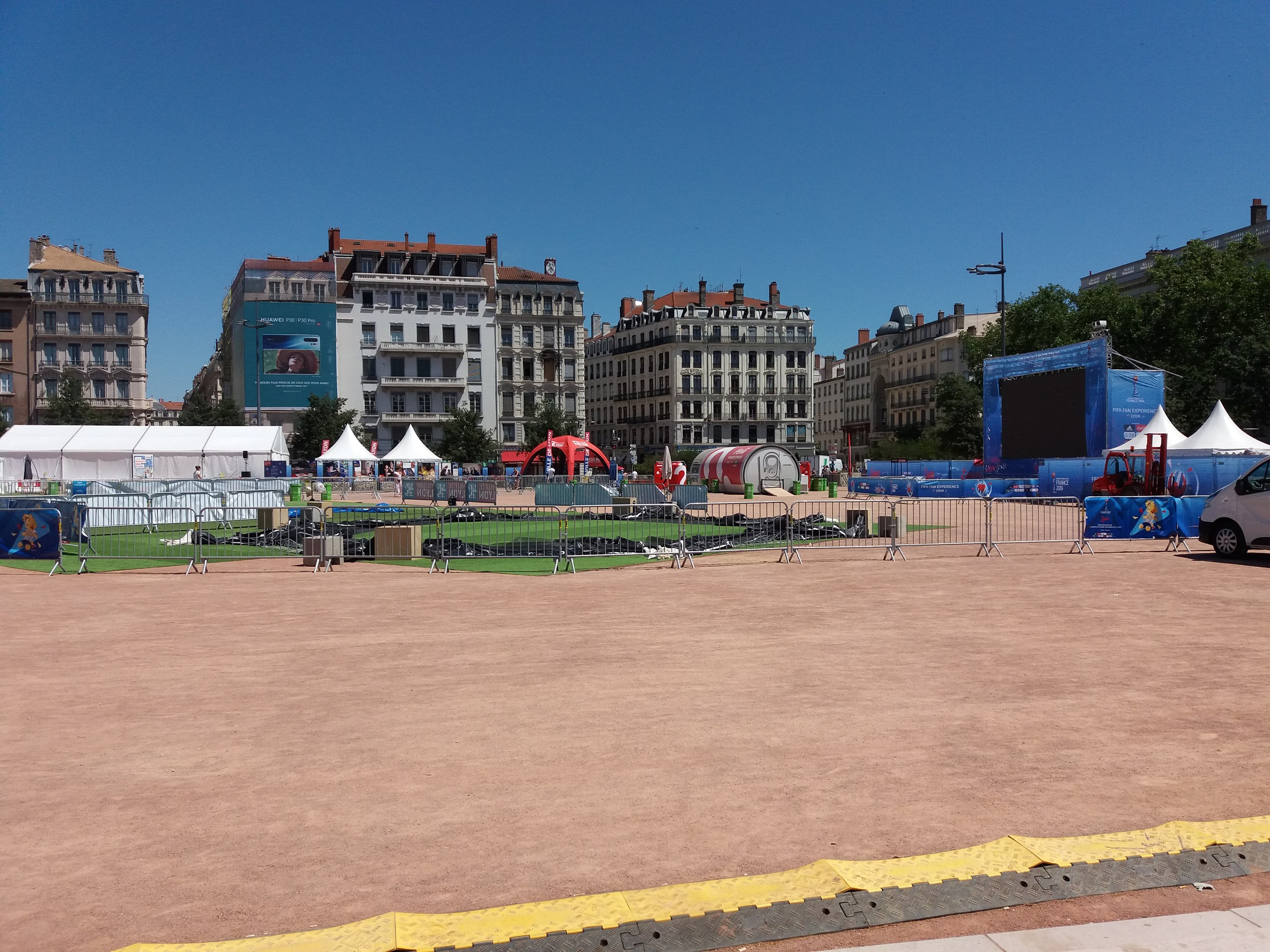 Großer Platz voller Zelte, umgeben von Gebäuden mit Fenstern, Bäumen, Fahrzeugen auf der Straße und einem Geländer unter einem klaren blauen Himmel.