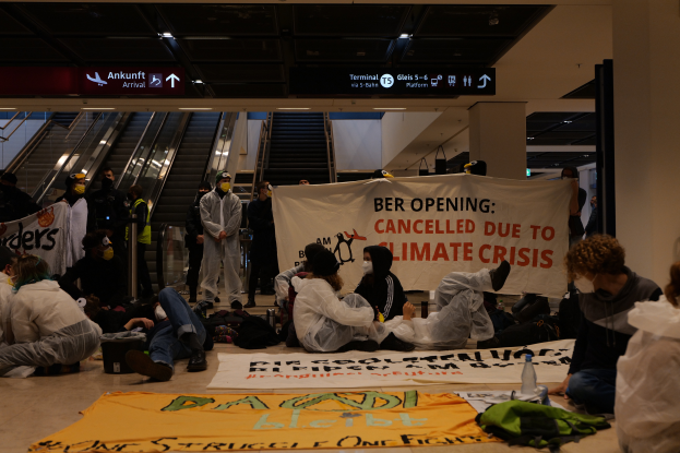 Eine Gruppe von Menschen sitzt auf dem Boden eines Flughafens und hält ein Banner in der Hand, auf dem "Berlin wegen Klimakrise gestrichen" steht. Um sie herum liegen verstreut Taschen, Flaschen und andere Gegenstände, und im Hintergrund sind Rolltreppen, Texttafeln und Deckenleuchten zu sehen.