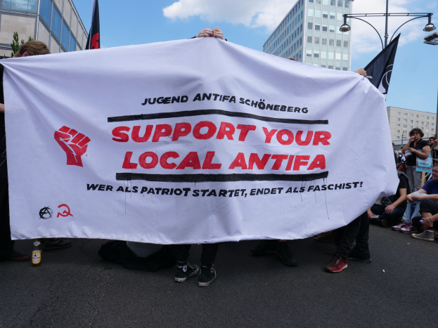 Eine Gruppe von Menschen auf einer Straße mit einer Fahne, auf der 'Support Your Local Antifa' und eine Flasche steht, und im Hintergrund Gebäude, Straßenmäste, Laternen, Bäume und ein bewölkter Himmel.