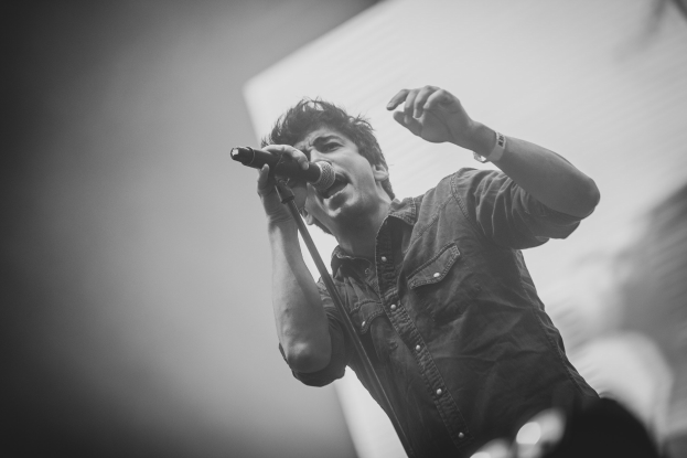 Schwarzes und weißes Foto eines Mannes, der in ein Mikrofon singt, bei einem Konzert mit einem unscharfen Gesicht im Hintergrund.