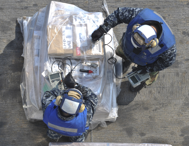 Zwei Männer in blauen Uniformen und Helmen arbeiten an einem großen Drohnenmodell, halten verschiedene Gegenstände mit einer Plastikabdeckung auf dem Boden.