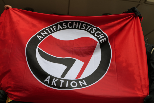 Eine Gruppe von Menschen mit einer roten Fahne mit der Aufschrift "Antifaschistische Aktion", die vor einer Wand steht.