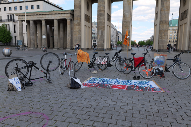 Eine Gruppe von Fahrr├Ądern, die vor dem Brandenburgertor in Berlin, Deutschland, geparkt sind, mit verstreuten Taschen und Gegenst├Ąnden, einer Fahne, Geb├Ąuden, B├Ąumen, Masten und einem klaren blauen Himmel im Hintergrund.