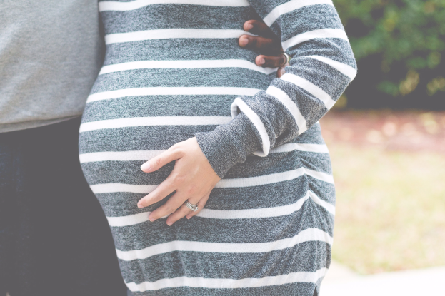 Schwangere Paare stehen nebeneinander mit den Händen auf dem Bauch, tragen Schwangerschaftskleidung in einem bewaldeten Gebiet.