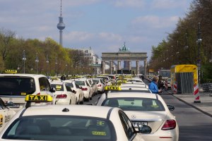 Eine belebte Straße in Berlin, Deutschland, mit zahlreichen geparkten Taxis, Fußgängern auf dem Gehweg, Laternenpfählen, Bäumen, Gebäuden und einem fernen Bogen mit Statuen und einem Turm unter einem bewölkten Himmel.