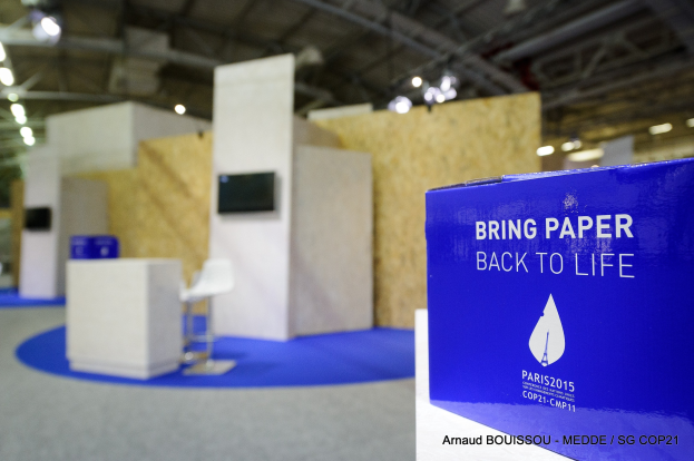 Messestand mit einem blauen und weißen "Bring Paper Back to Life"-Schild auf der rechten Seite, der einen unscharfen Stuhl, Tische, einen wandmontierten Fernseher und Deckenleuchten im Hintergrund zeigt.