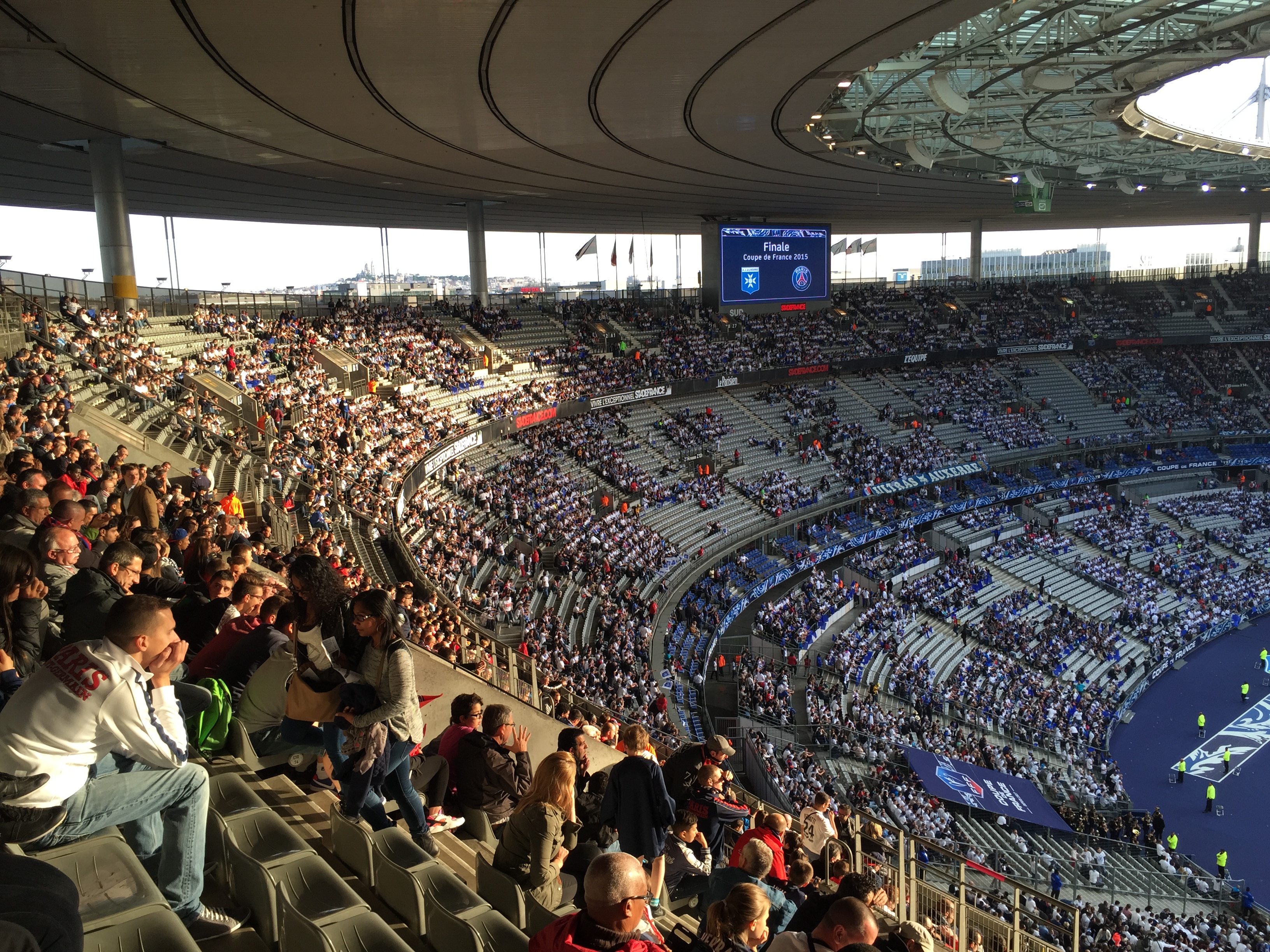 Eine große Menschenmenge sitzt im Allianz Stadion in München, Deutschland, bei einem Fußballspiel, mit einer Bühne auf der rechten Seite, Fahnen, Stangen und einem Bildschirm im Hintergrund und der Himmel oben sichtbar.