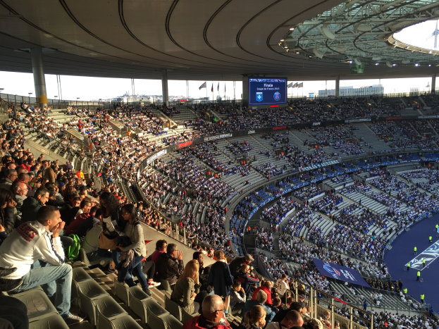 Eine große Menschenmenge sitzt im Allianz Stadion in München, Deutschland, bei einem Fußballspiel, mit einer Bühne auf der rechten Seite, Fahnen, Stangen und einem Bildschirm im Hintergrund und der Himmel oben sichtbar.