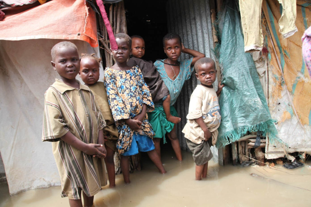 Eine Gruppe von Kindern steht vor einer Hütte, die von Wasser umgeben ist, mit Kleidung, die an einer Leine auf der rechten Seite hängt und einer Wand auf der linken Seite, ihre Gesichter zeigen Trauer und Verzweiflung.