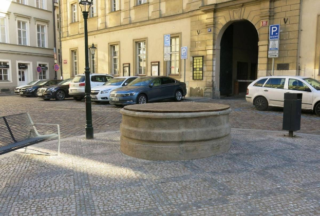 Ein Stadtplatz mit einer Bank, einem Brunnen, Gebäuden mit Fenstern, Laternen und Schildern.