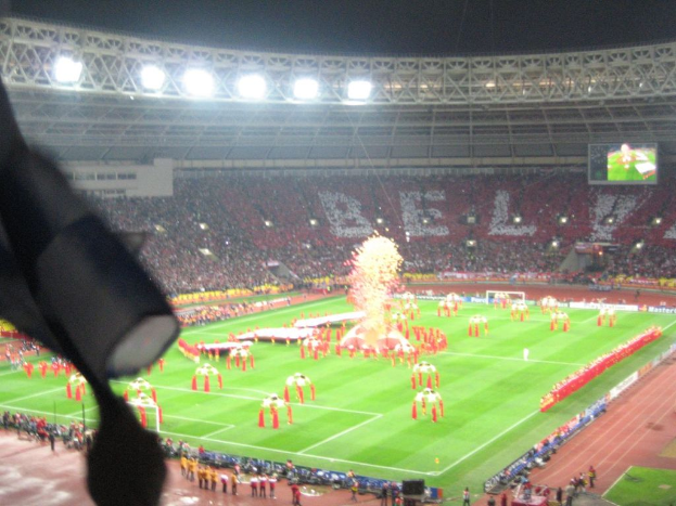 Ein Stadion mit Flutlicht, Eisenstäbe, Zuschauern, stehenden Personen, Werbetafeln, Anzeigetafeln, Überwachungskameras und dekorativen Fahnen.