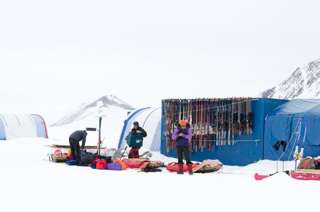 Drei Personen stehen auf einer verschneiten Landschaft mit verstreuten Taschen, Zelten mit Skiern darauf und schneebedeckten Hügeln im Hintergrund unter einem klaren Himmel.