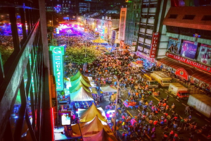 Ein buntes Karnevalsszenario mit einer großen Menschenmenge auf der Straße, umgeben von Zelten, Geschäften, Gebäuden und Bannern, mit zahlreichen Lichtern im Hintergrund.
