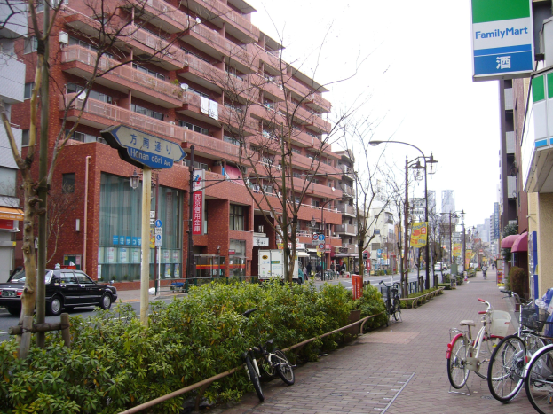 Stadtstraße mit parkenden Fahrrädern, Gebäuden, Straßenlaternen, Schildern, fahrenden Fahrzeugen, Fußgängern auf dem Gehweg, Grünflächen und einem klaren blauen Himmel.