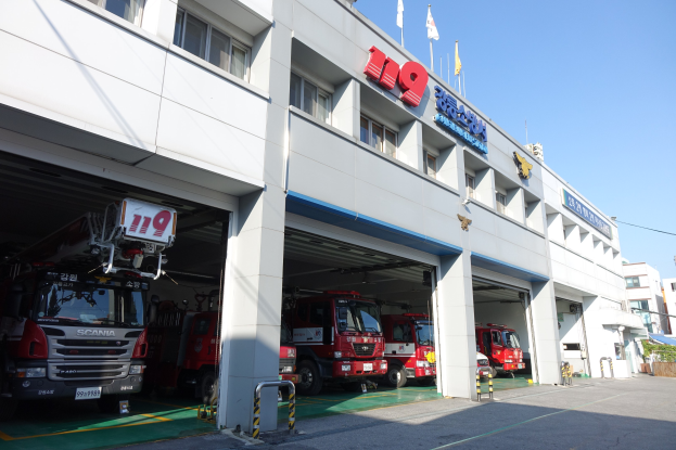 Feuerwache mit mehreren Feuerwehrautos davor, mit Fenstern, Säulen und Namensschildern, sowie Flaggen im Vordergrund und anderen Gebäuden gegen einen klaren blauen Himmel.