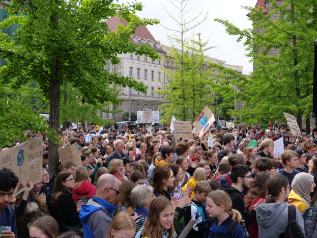 Eine Menschenmenge mit Schildern steht vor einem Gebäude, Bäume, Fahrzeuge und ein Redner sind in Berlin zu sehen.