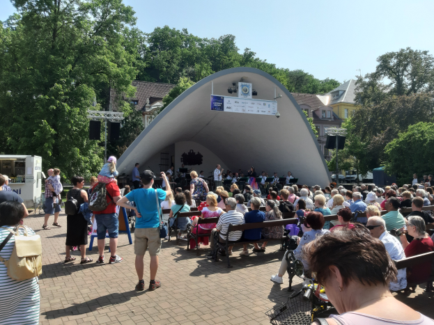 Eine Menschenmenge sitzt auf Bänken und steht vor einer Bühne auf einem Sommerkonzert im Park, mit einer Person mit einer Kamera, einem Gerät auf einem Ständer, einer Fahne mit Text, Pfosten, einem Metallrahmen, Bäumen, Gebäuden mit Fenstern und einem bewölkten Himmel.