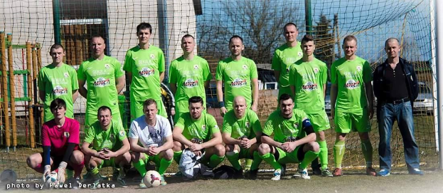 Eine Gruppe von Männern in grünen T-Shirts steht auf einem Fussballfeld in der Nähe eines Torpfostens, mit einem Ball vor ihnen und verschiedenen Hintergrundelementen wie Bäume und Gebäude.